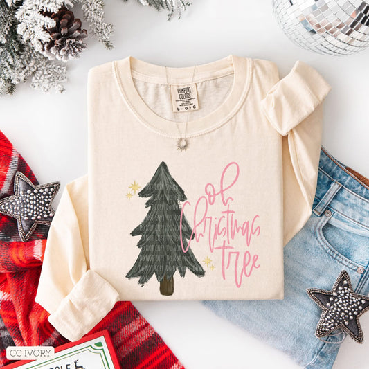 Beige t-shirt with Christmas tree design and text, surrounded by festive items on a white surface.