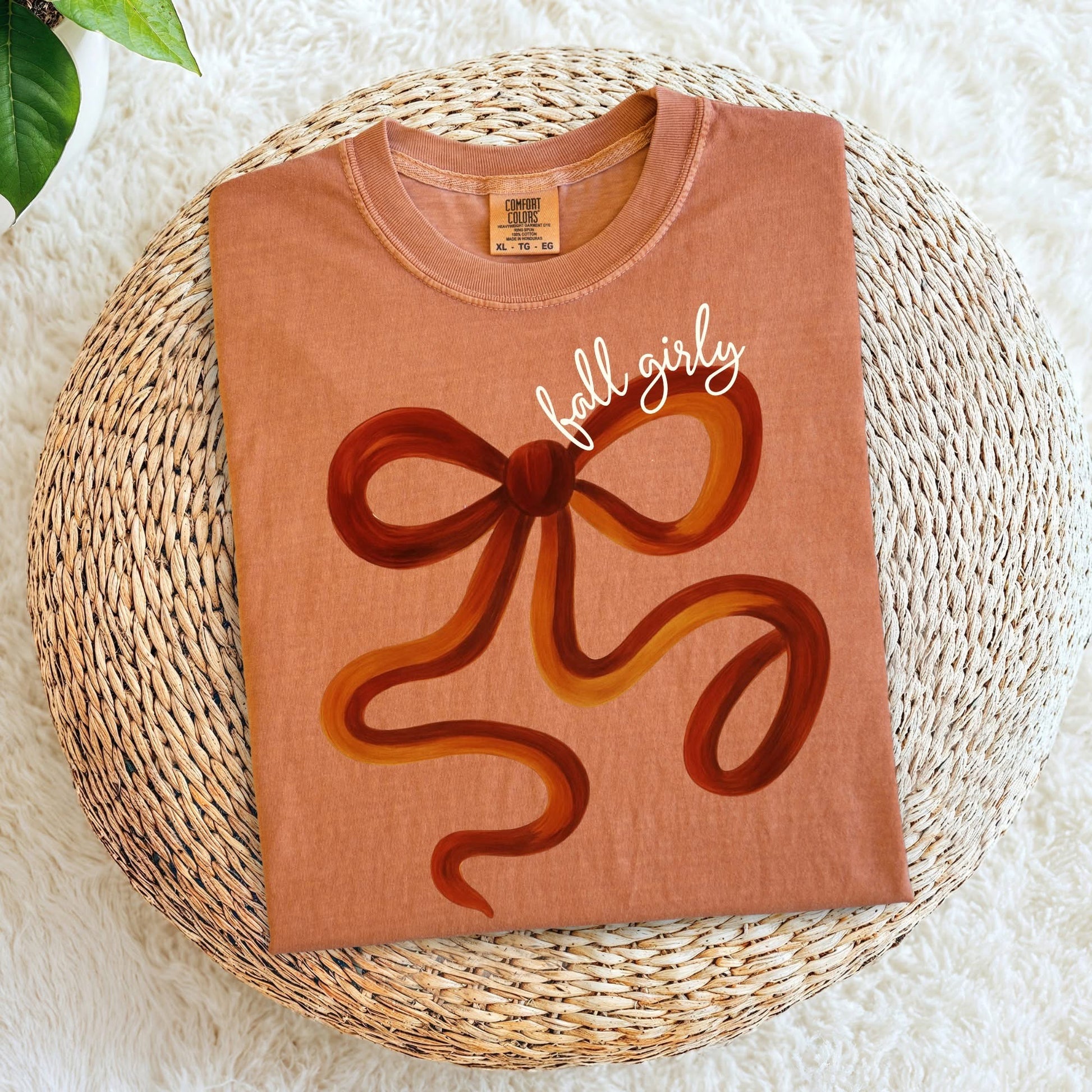 Brown t-shirt with a large brown bow graphic and 'fall girly' text on a woven mat.