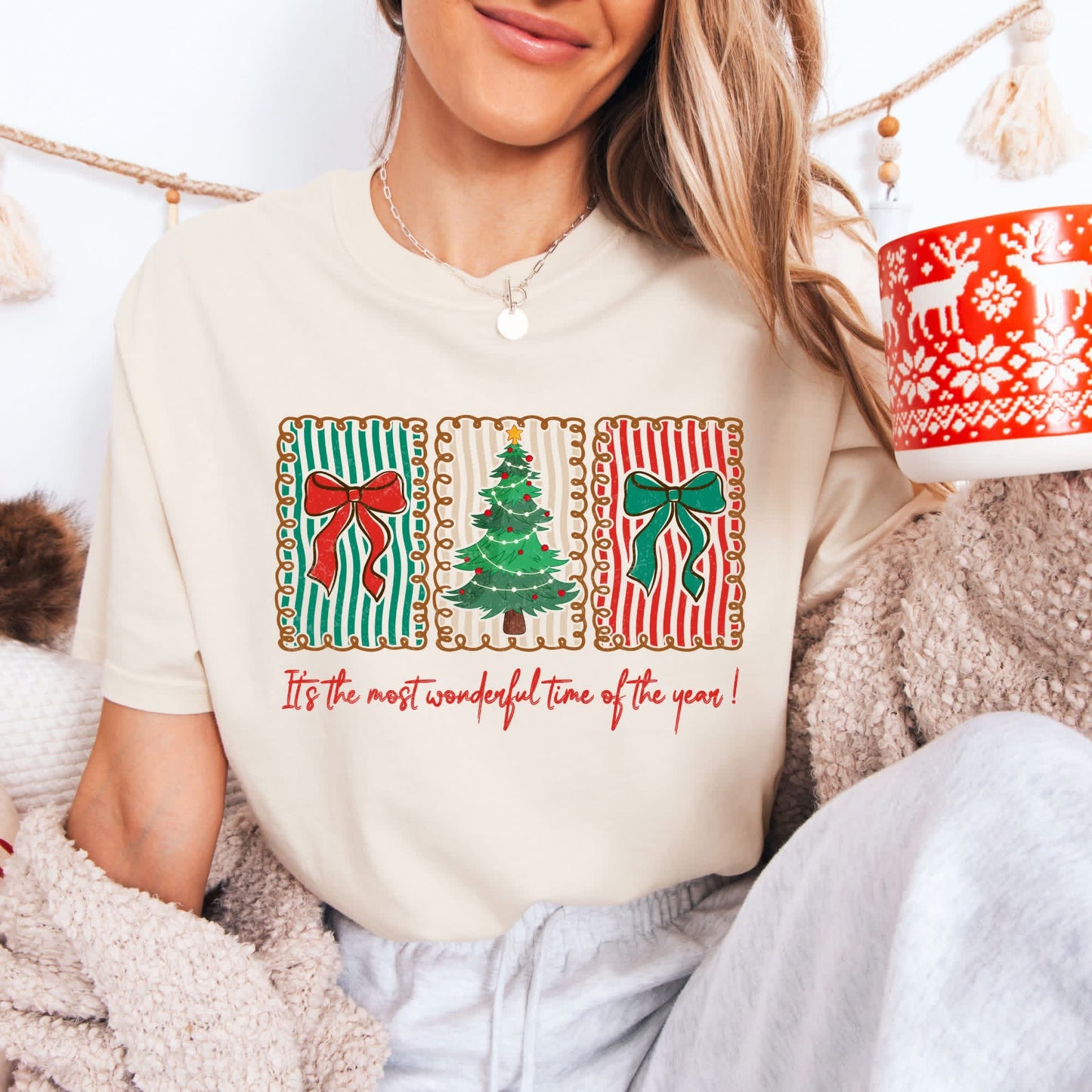 Person wearing a cream-colored t-shirt with Christmas-themed graphics and text, sitting in a cozy setting.