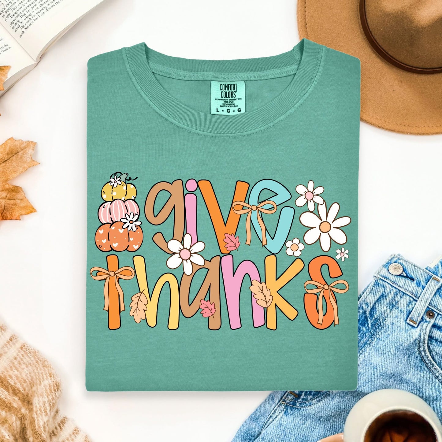 Green t-shirt with 'Give Thanks' text and fall-themed graphics on a white surface with accessories.