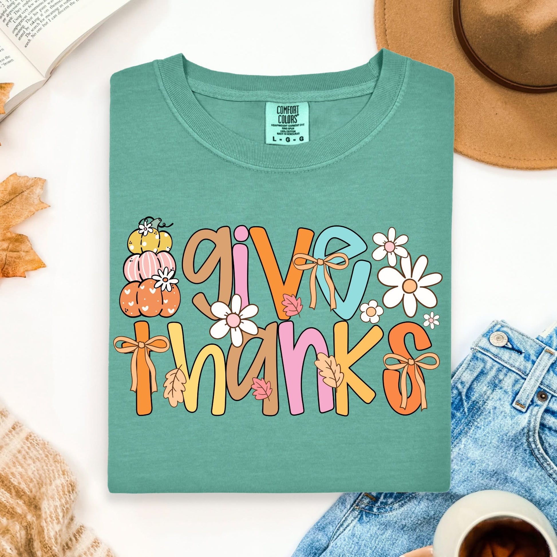 Green t-shirt with 'Give Thanks' text and fall-themed graphics on a white surface with accessories.