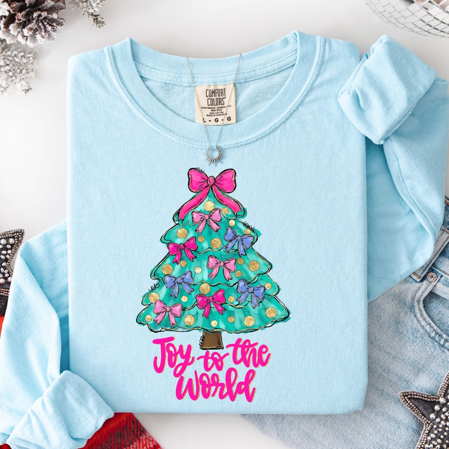 Light blue long sleeve shirt with a Christmas tree design and 'Joy to the World' text on a white surface.