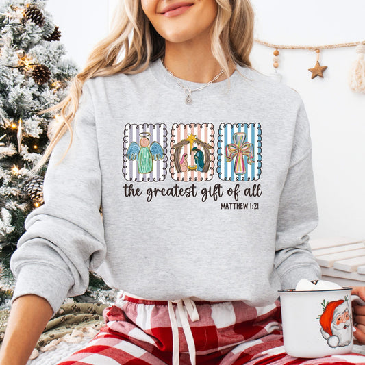 Person wearing a gray sweatshirt with angel, nativity, and cross designs and text, sitting in front of a Christmas tree.