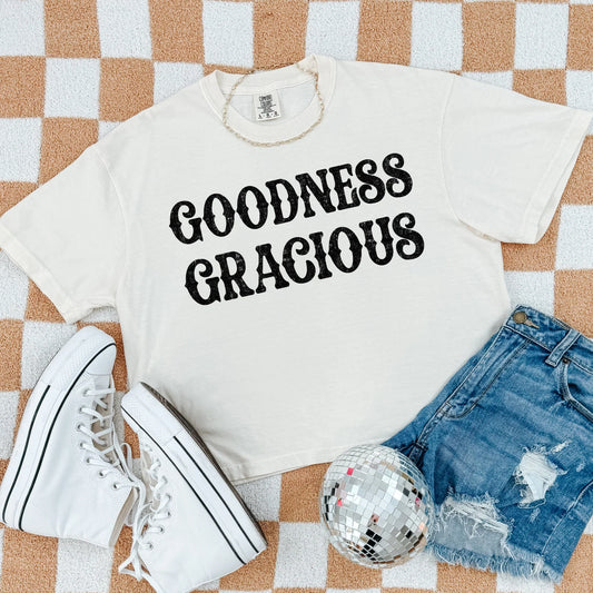 White t-shirt with 'GOODNESS GRACIOUS' text, white sneakers, denim shorts, and disco ball on a checkered background