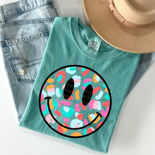Teal t-shirt with colorful smiley face design on a white surface with denim jeans and a hat.