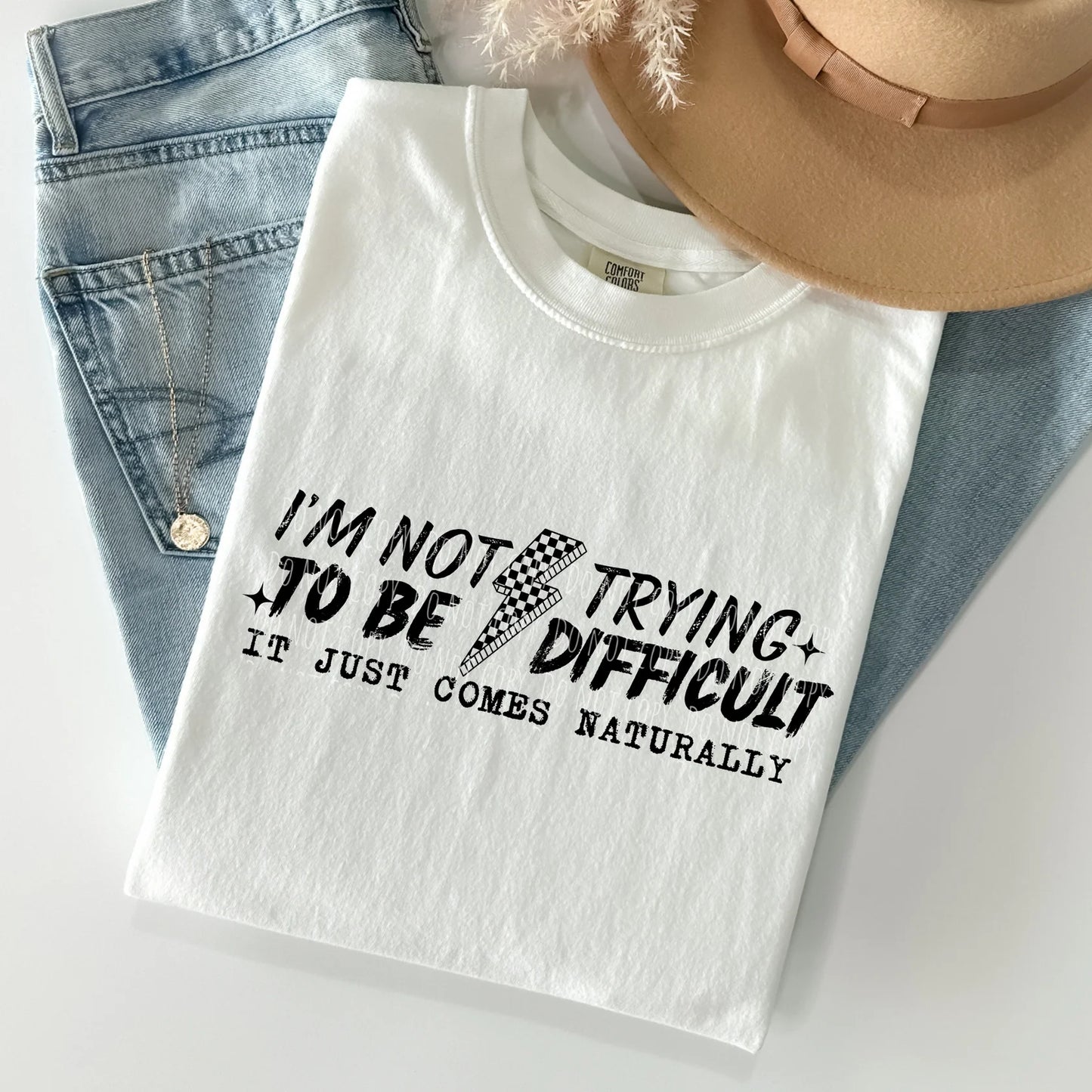 White t-shirt with humorous text design on a light gray surface with jeans and a hat in the background.