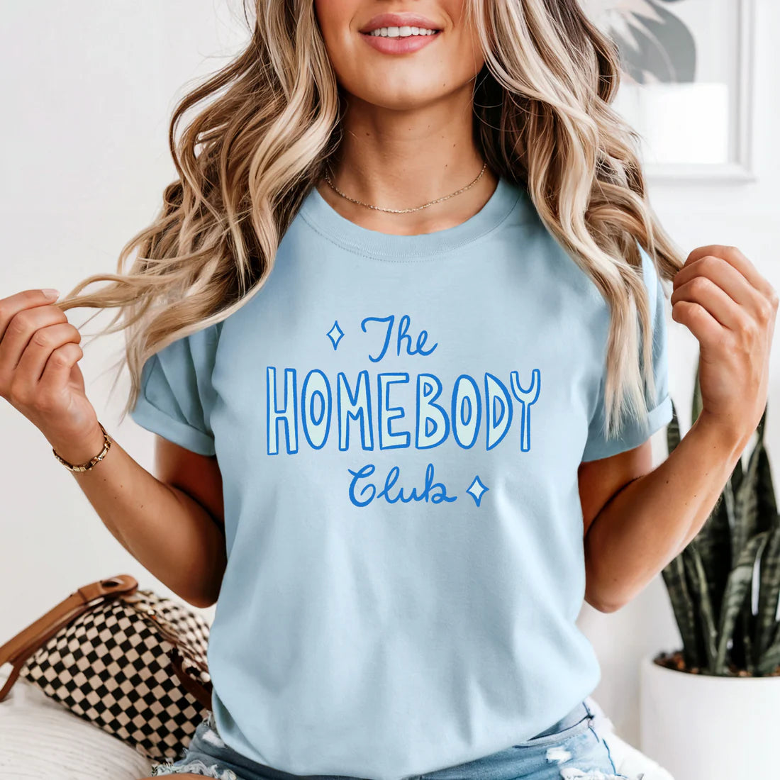 Woman wearing a light blue t-shirt with 'The Homebody Club' text.