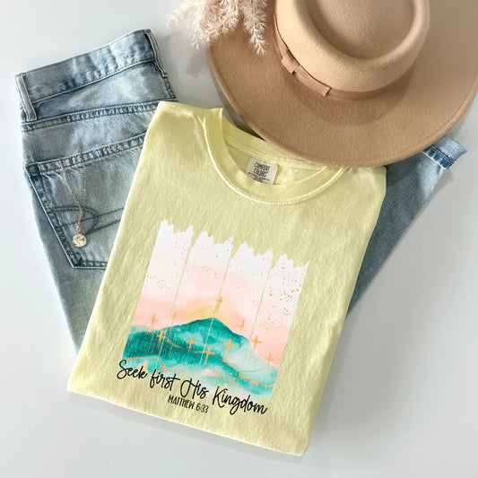 Yellow t-shirt with a scenic design and text, placed on denim jeans and a hat.
