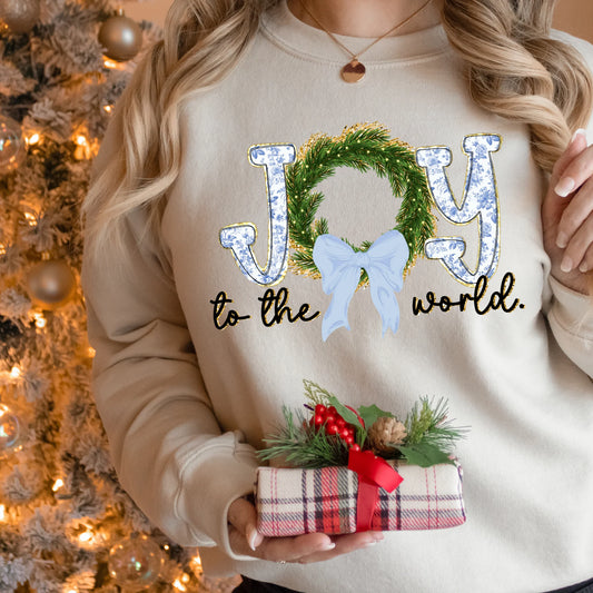 Person wearing a beige sweatshirt with blue toile 'Joy to the world' text, holding a small gift, Christmas tree in the background