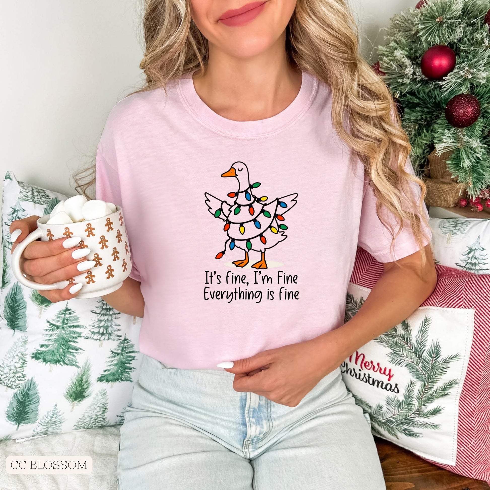 Female with blonde hair wearing a pink t-shirt with a Christmas design of white goose tangled in colored Christmas lights. Model is holding a mug, and sitting in front of Christmas pillows and small decorated Christmas tree.