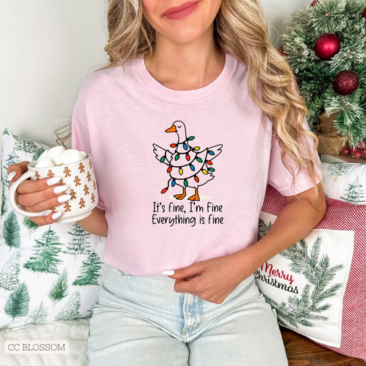 Female with blonde hair wearing a pink t-shirt with a Christmas design of white goose tangled in colored Christmas lights. Model is holding a mug, and sitting in front of Christmas pillows and small decorated Christmas tree.