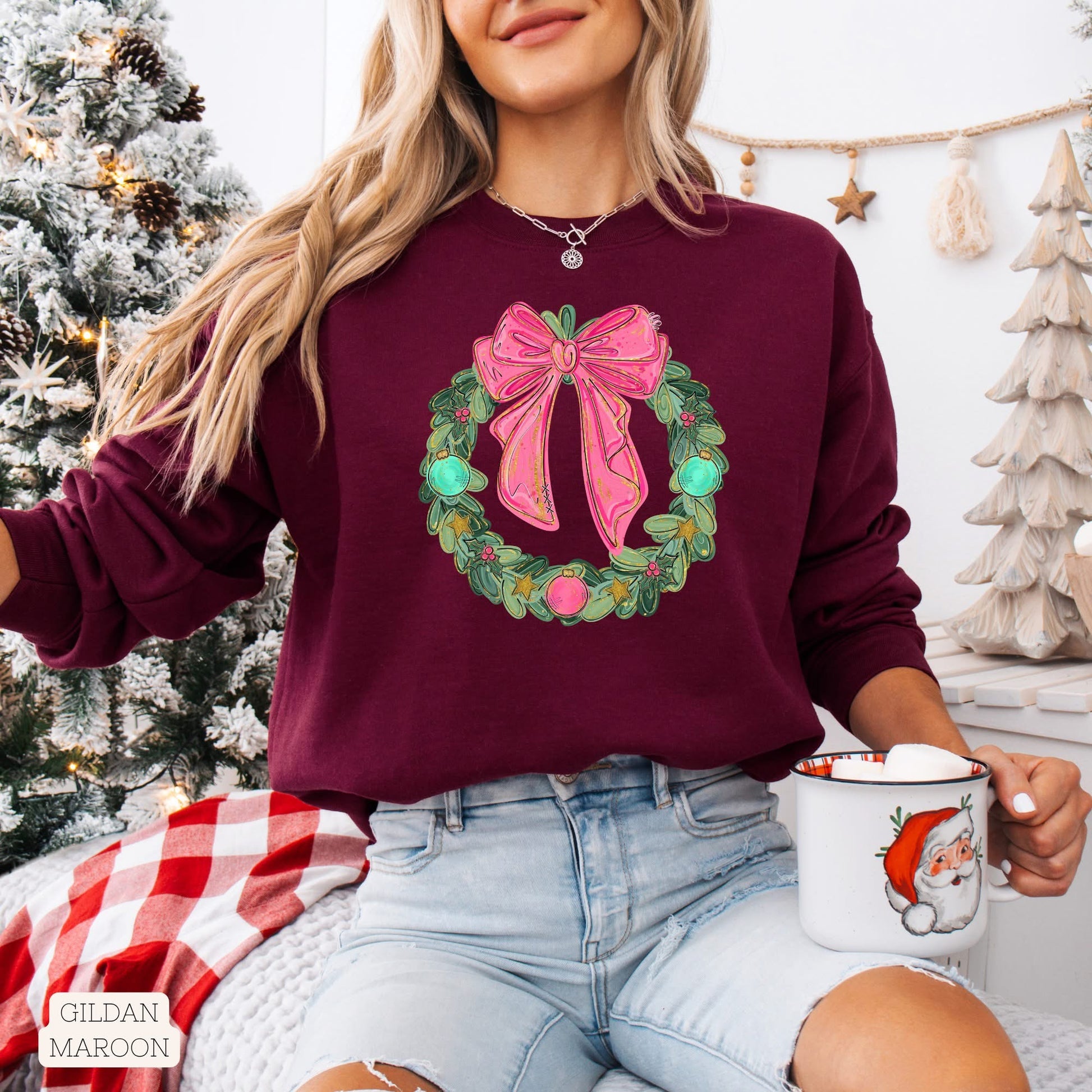 Female with blonde hair wearing a maroon long sleeve tee with a Christmas wreath design with big pink bow, holding a mug in a festive setting.