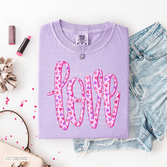 Folded purple shirt, with handwritten word love with shades of pink, red, and purple hearts on the don’t, laying on cut off blue jean shorts on a white background with lipstick